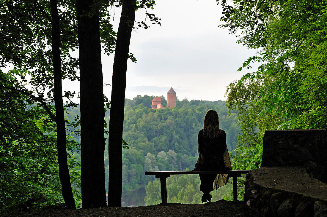  Frau sitzt auf einer Bank mit Blick auf den Fluss Gauja mit der Burg Turaida im Hintergrund, rund um Sigulda, Gauja-Nationalpark, Region Vidzeme, Lettland, Baltikum, Nordeuropa 