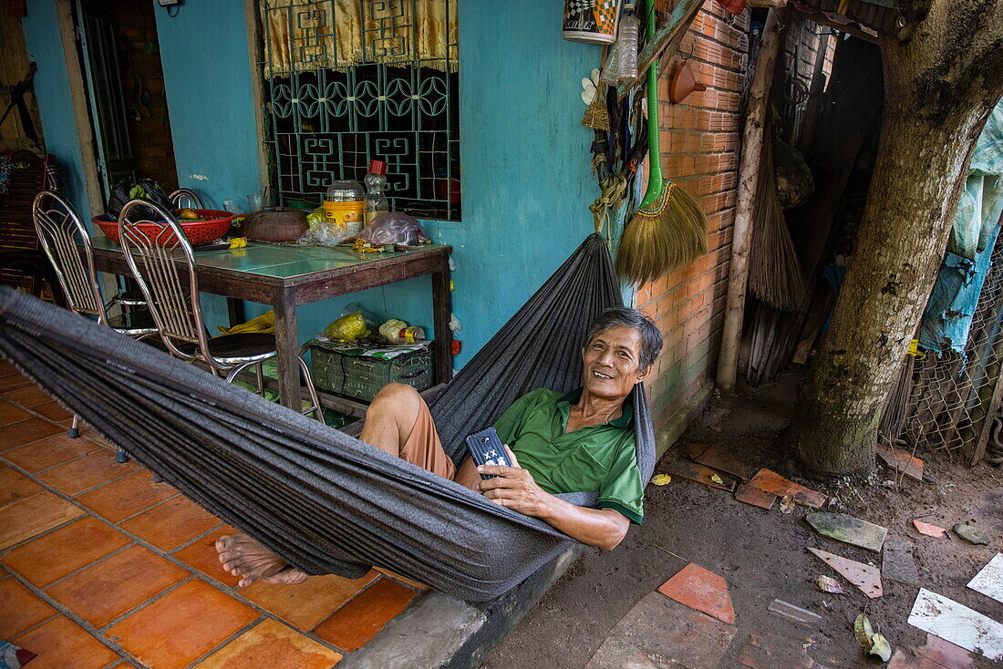  Man relaxing in hammock and smiling while looking up from his smartphone, Long Son, Phu Tan, An Giang, Vietnam, Asia 