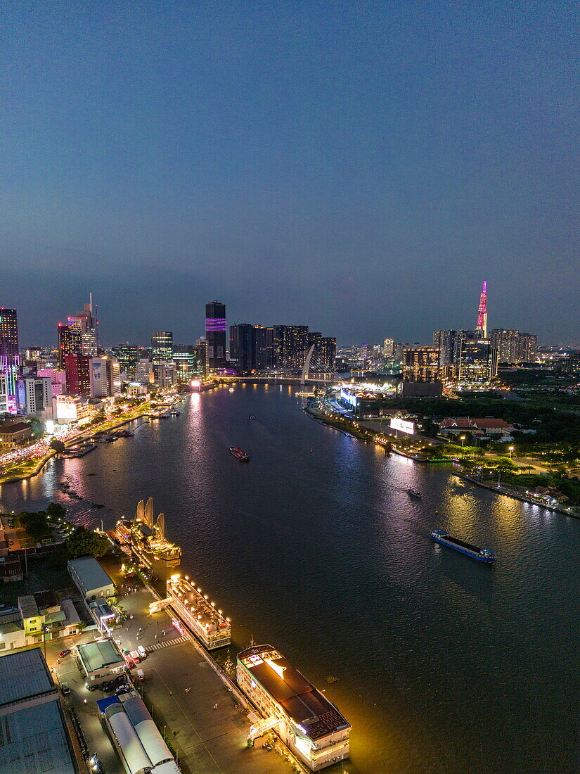  Aerial view of illuminated tourist tour boats along Saigon River with skyscrapers in District 1 city center and Landmark 81 tower in the distance at dusk, Quan 4, Ho Chi Minh City, Vietnam, Asia 