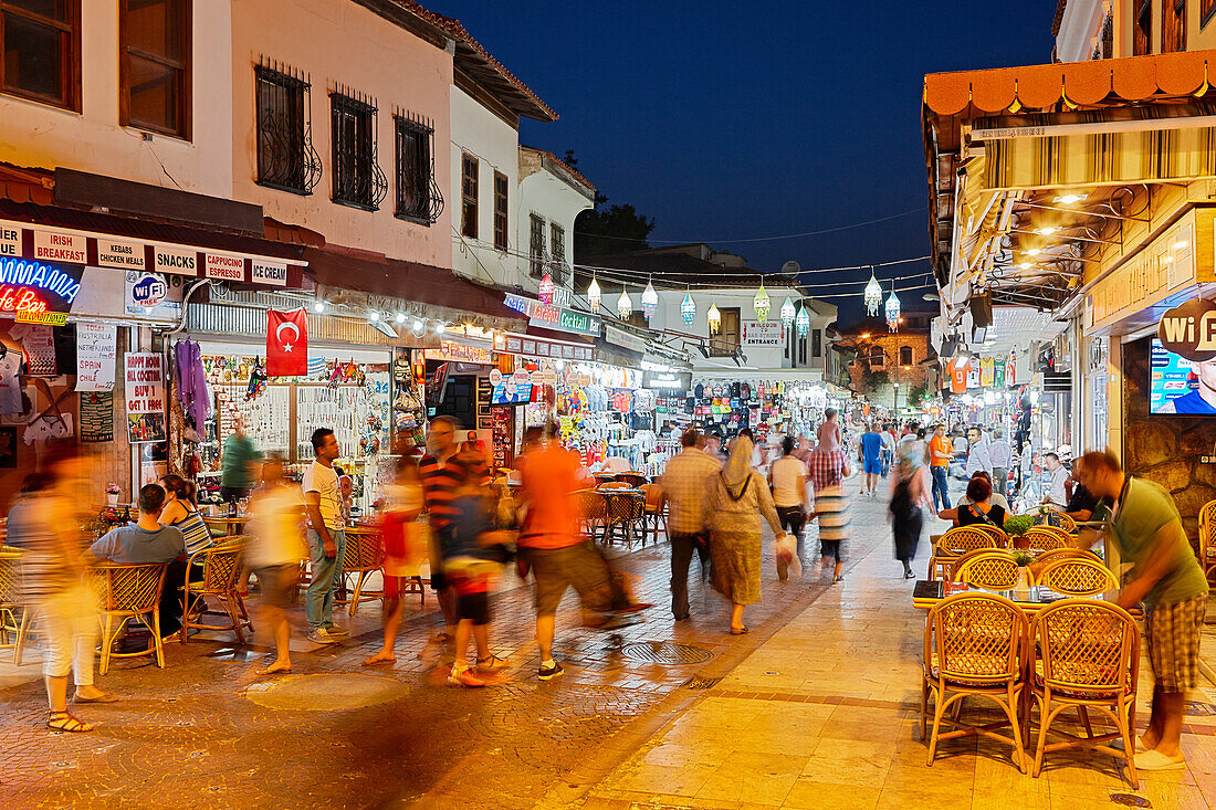 People walk in a narrow street brightly illuminated at night in Kusadasi, a large resort town on the Aegean coast of Aydin Province, Turkey.