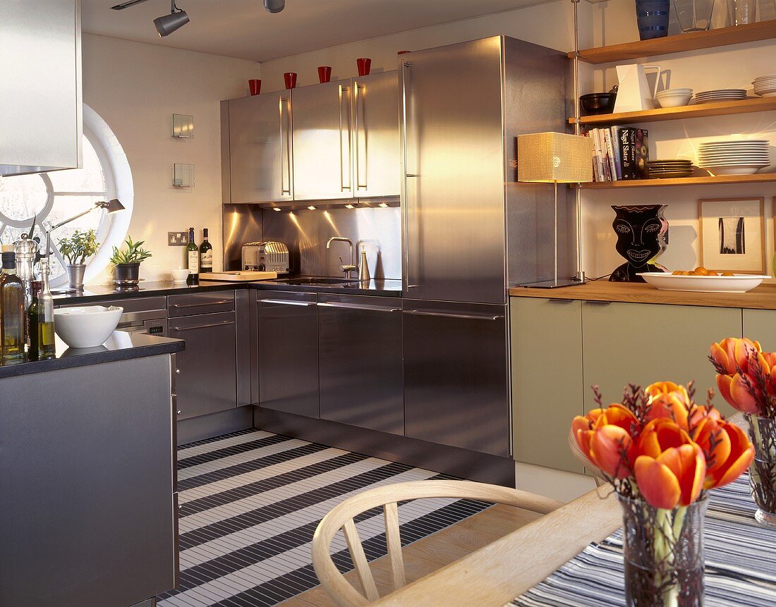 An open-plan kitchen with stainless steel cupboards and a wooden shelf stacked with crockery