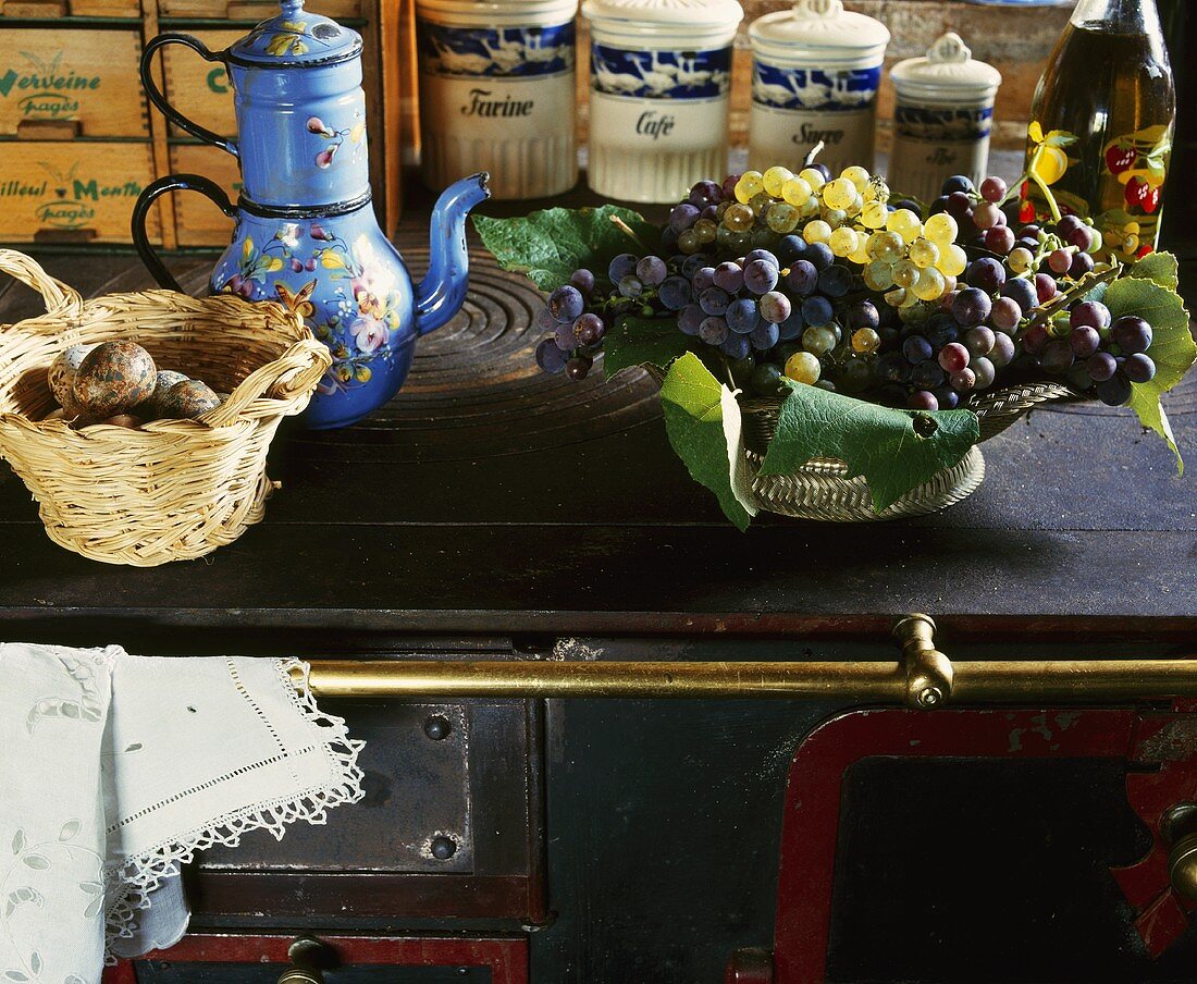 Eine alte blaue Emaille-Kaffeekanne, ein Korb mit Fasaneneiern und eine Schale mit Trauben auf einem alten Herd