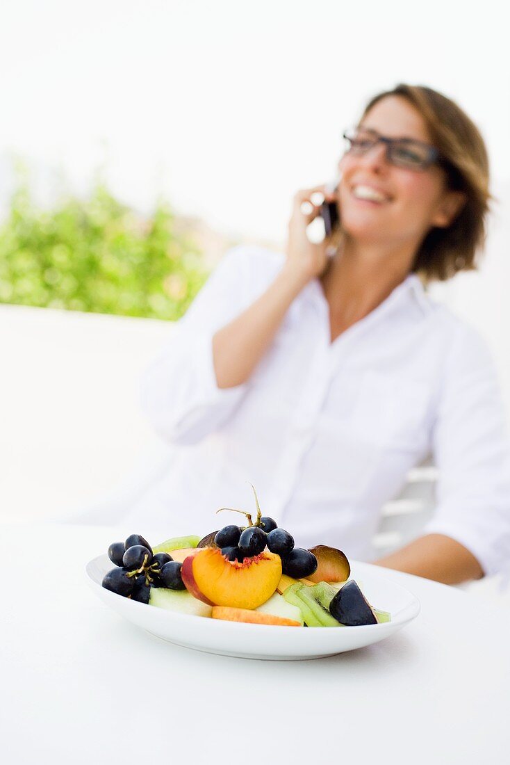 Obstteller, dahinter Frau mit Mobiltelefon