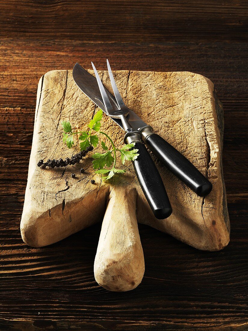 A chopping board with a carving knife and fork, parsley and peppercorns