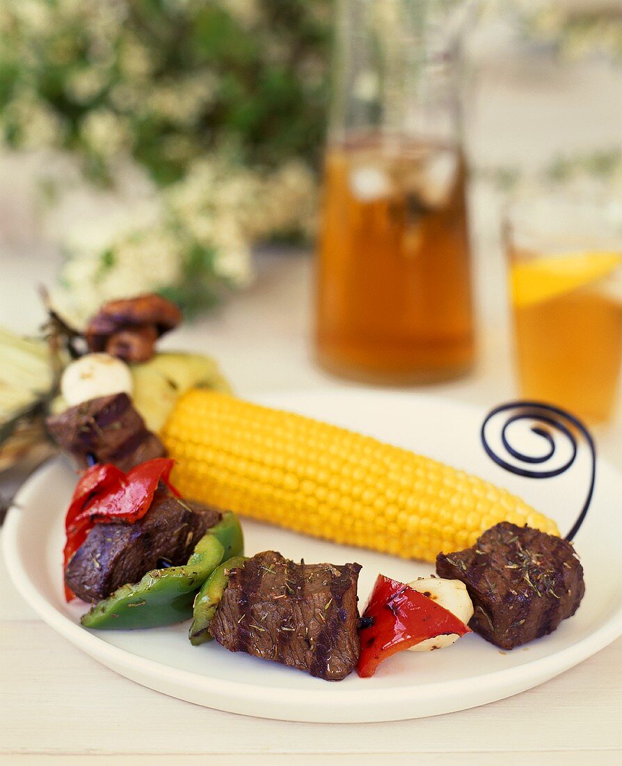 Barbecued meat kebabs with corncobs; iced tea