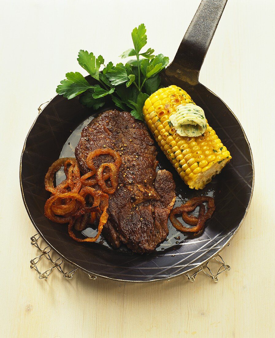 Beef steak with onion rings and corncob in frying pan