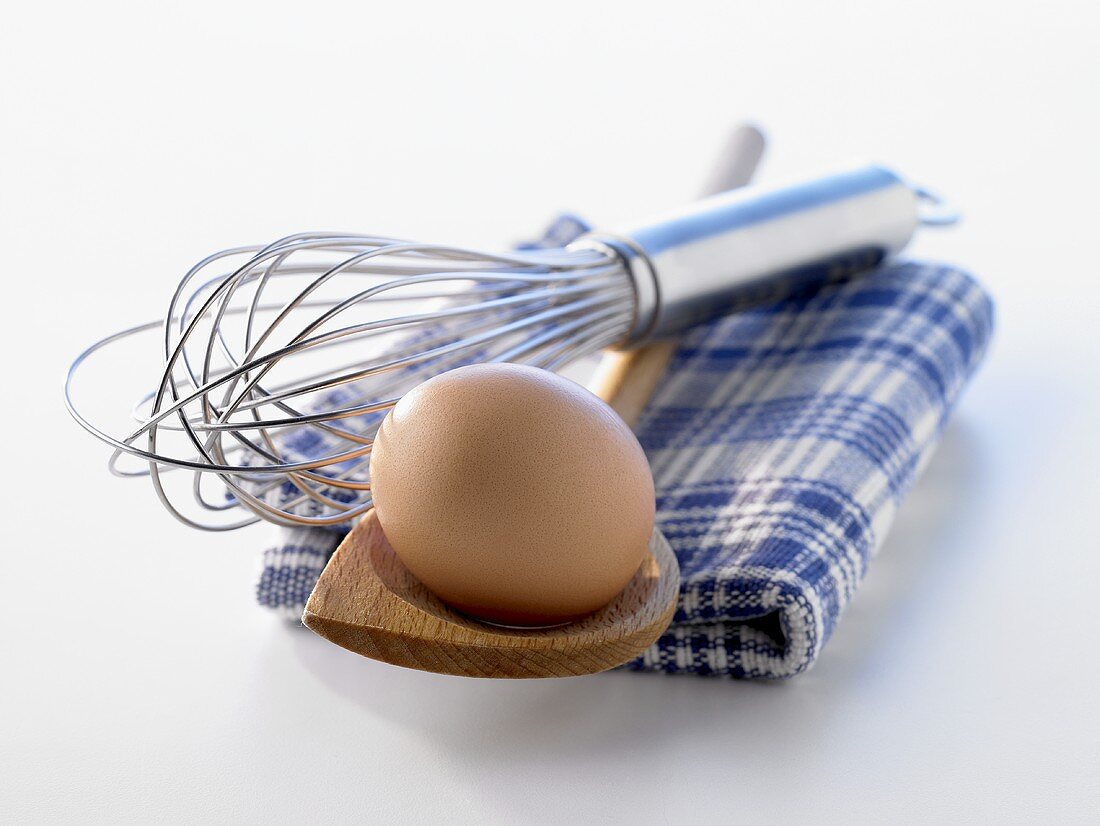 A brown egg on wooden spoon with egg whisk & kitchen cloth