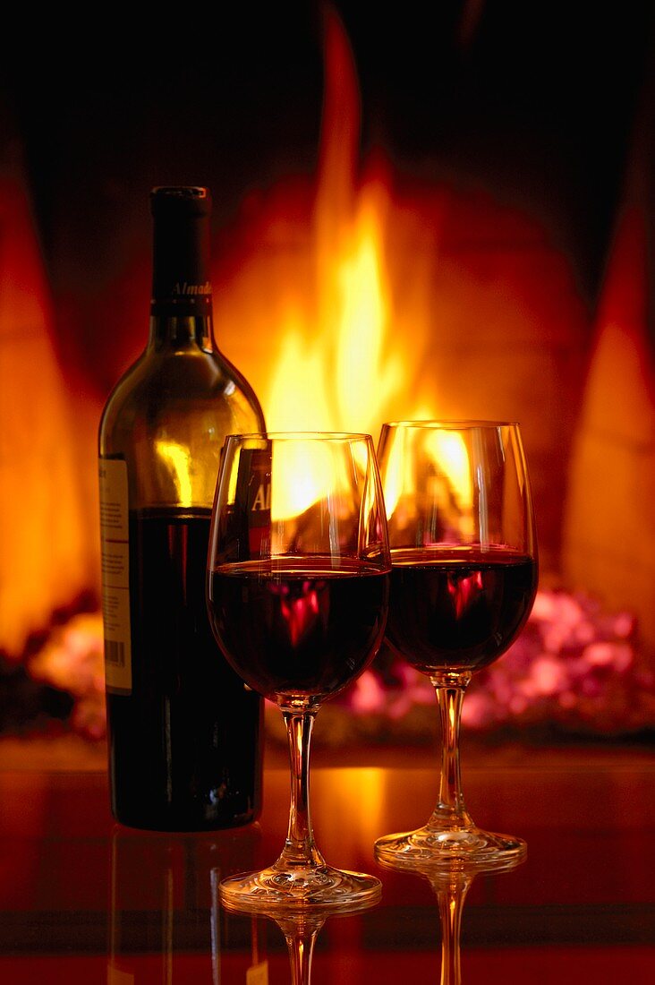 Eine Flasche Rotwein mit zwei Gläsern vor Kamin-Feuer