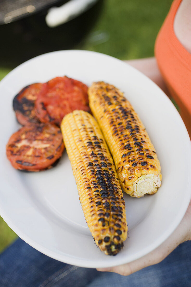 Woman holding plate of grilled corn cobs and tomatoes