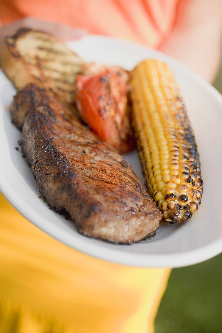 Woman holding plate of grilled beef steak & corn on the cob