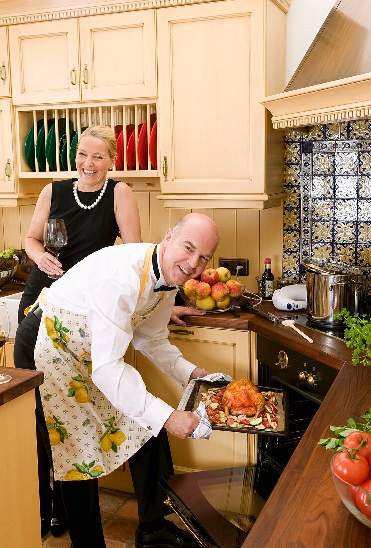 Man putting duck into oven, woman behind with glass of red wine