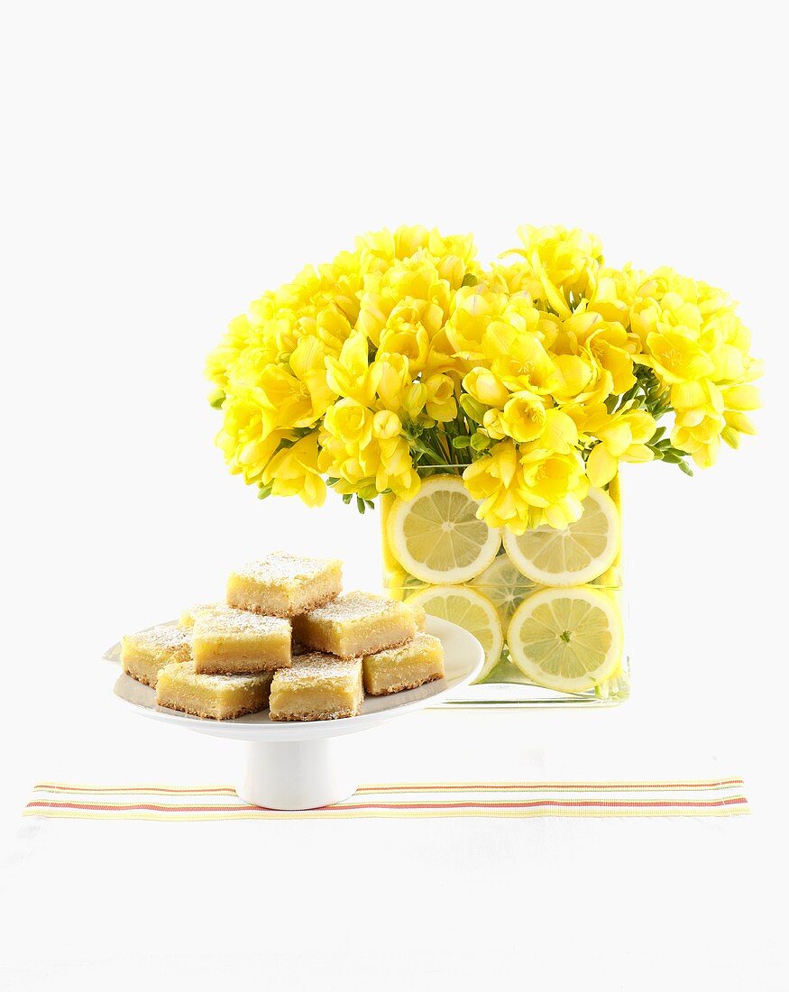 Zitronenschnitte auf Kuchenständer und gelbe Fresien