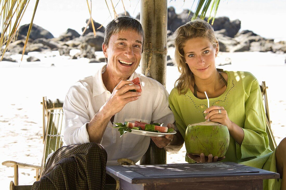 Father eating watermelon, daughter drinking coconut milk on beach