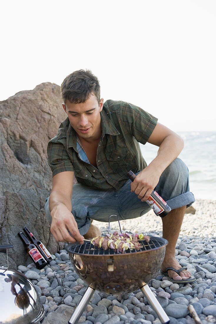 Junger Mann wendet Schaschlik-Spiesse auf Grill am Strand