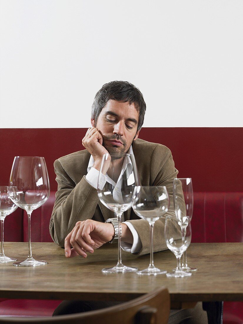 Mann sitzt mit leeren Weingläsern am Tisch und schaut auf seine Uhr
