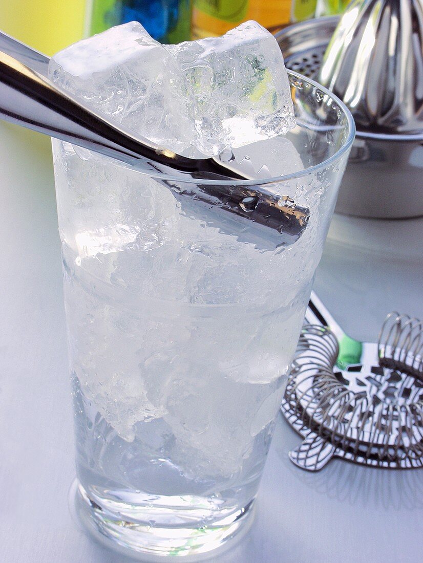 Glass with ice cubes & ice tongs in front of bar utensils