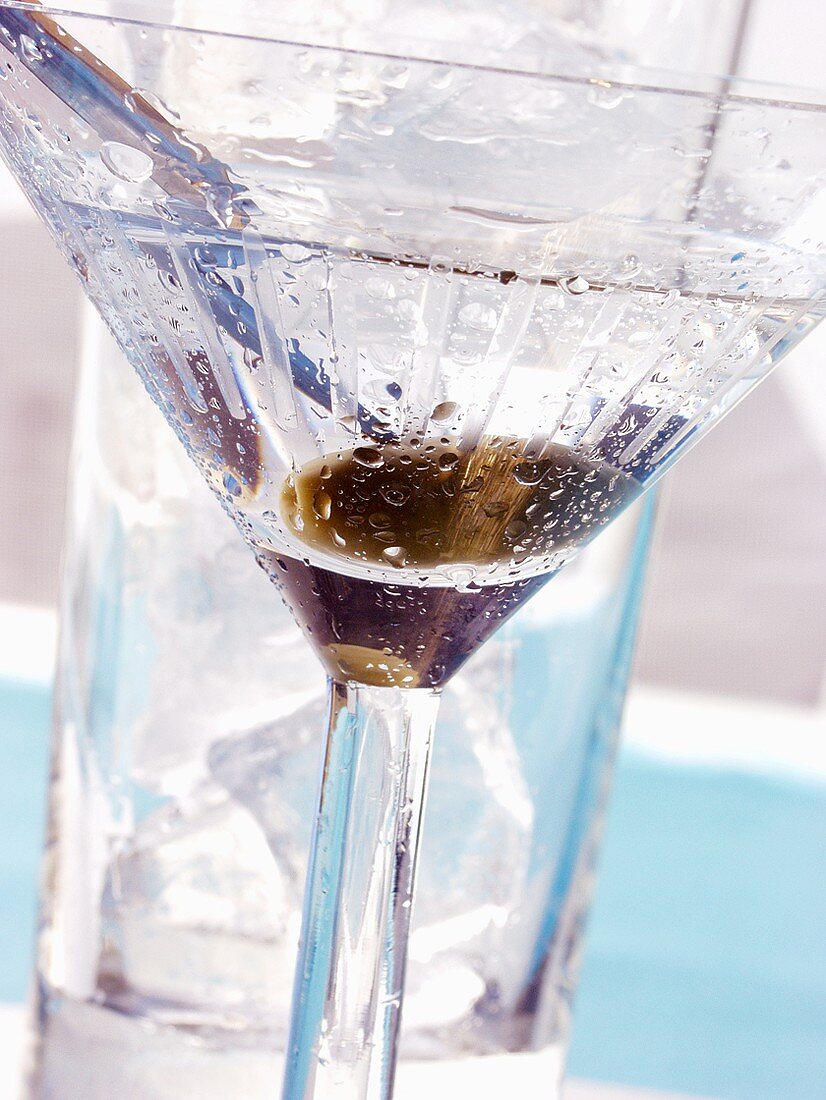 Martini with green olive in front of glass with ice cubes