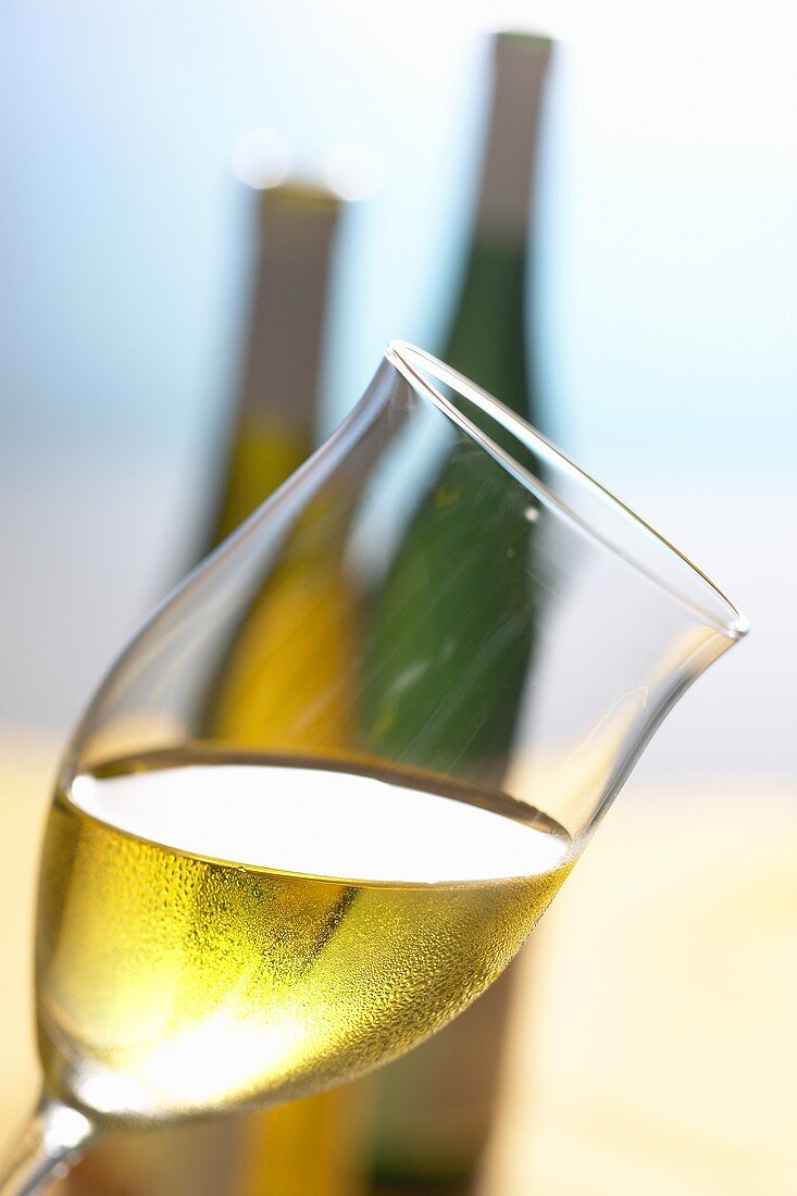 Glass of white wine, held at an angle, in front of two bottles