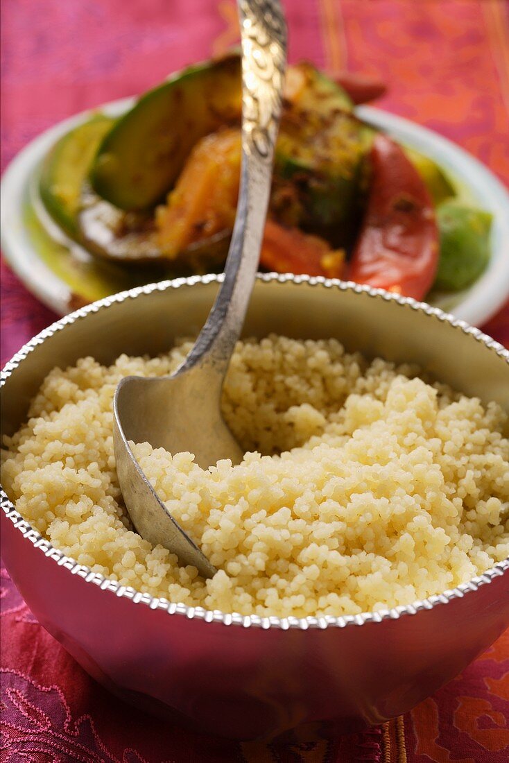 Couscous in silver bowl, plate of vegetables behind