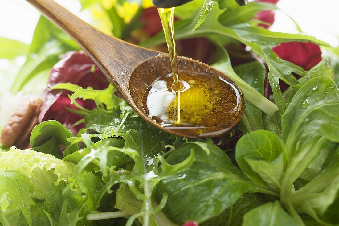 Pouring olive oil into wooden spoon above salad leaves