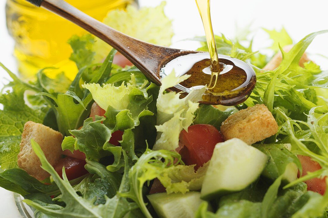 Pouring olive oil into wooden spoon above salad leaves