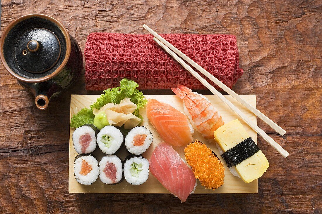 Verschiedene Sushi auf Sushibrett, daneben Tuch und Kännchen