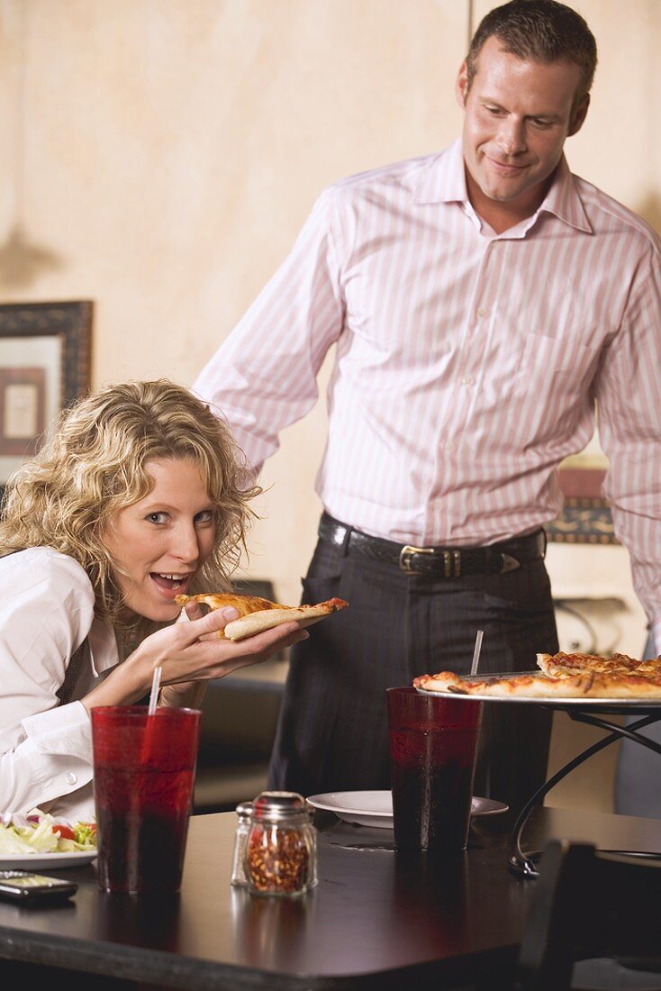 Woman in restaurant eating pizza, man just arriving