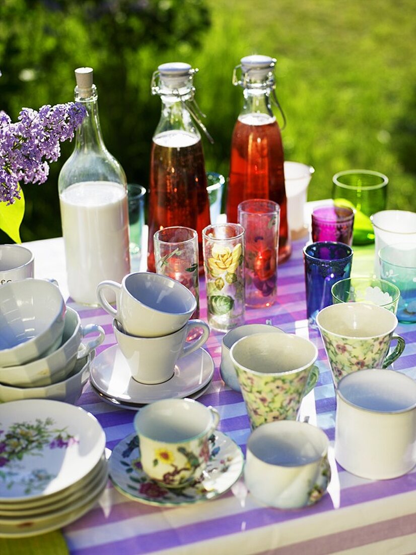 Tisch mit Tassen, Gläsern, Getränken & lila Flieder im Freien