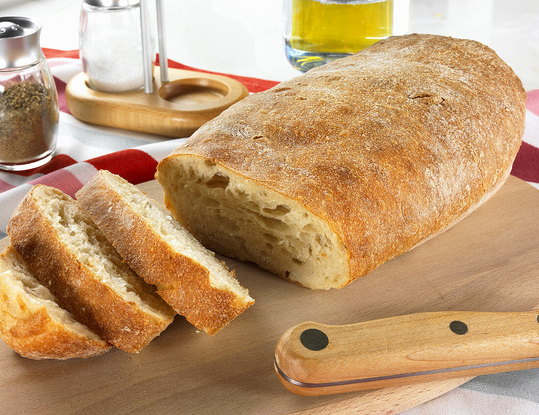 Ciabatta auf Holzplatte, dahinter Pfeffer, Salz und Olivenöl