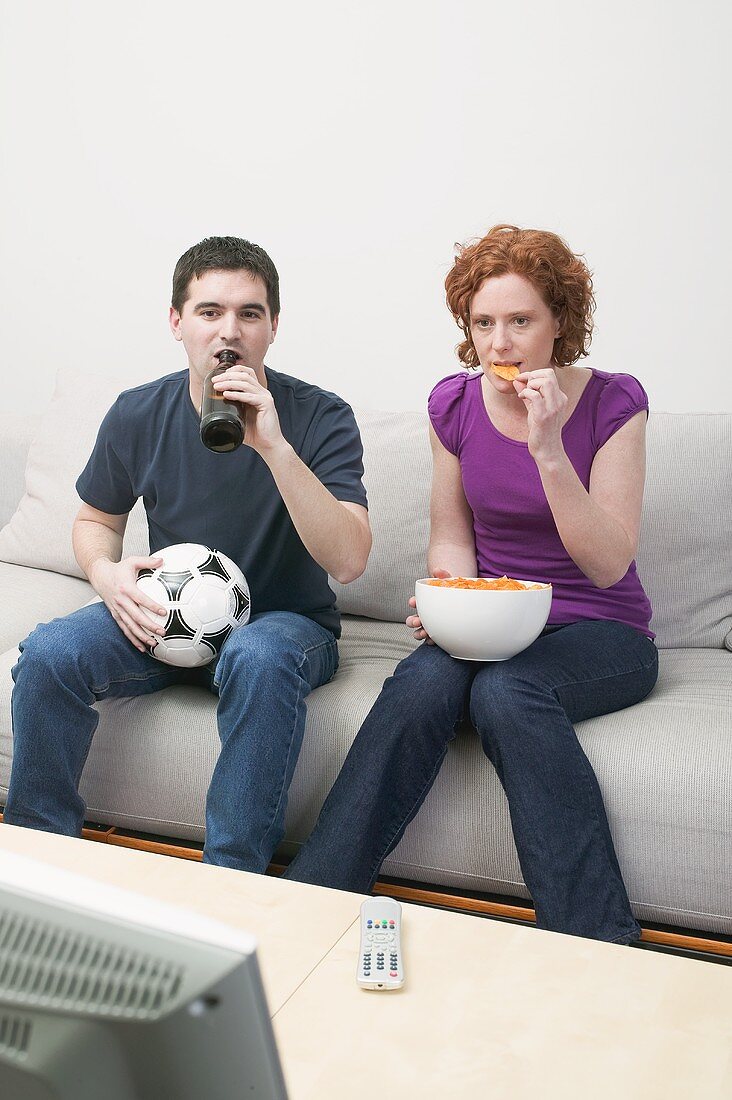Junges Paar mit Fussball, Bierflasche & Chips beim Fernsehen
