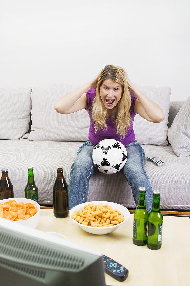 Young woman with football, beer bottles & crisps watching TV