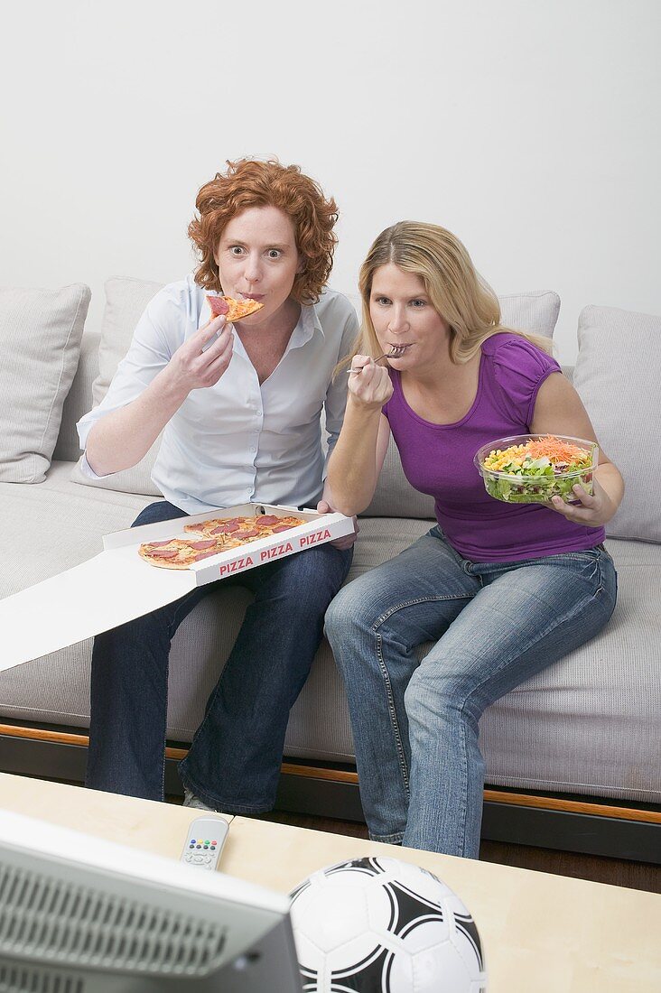 Two friends eating pizza and salad while watching TV