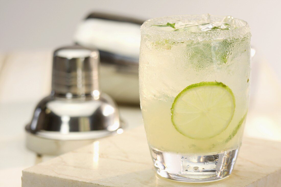 A mojito in a glass in front of a cocktail shaker