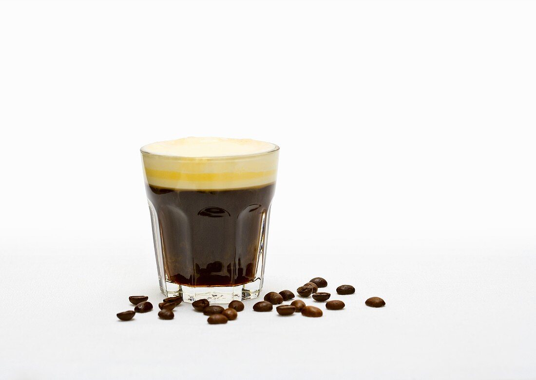 Coffee with cream in a glass with coffee beans next to it