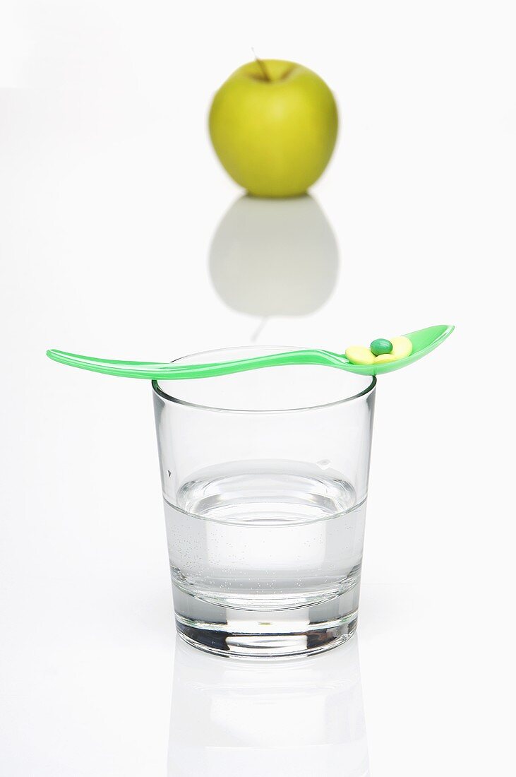 A vitamin tablet on a spoon on top of a glass of water with a green apple in the background