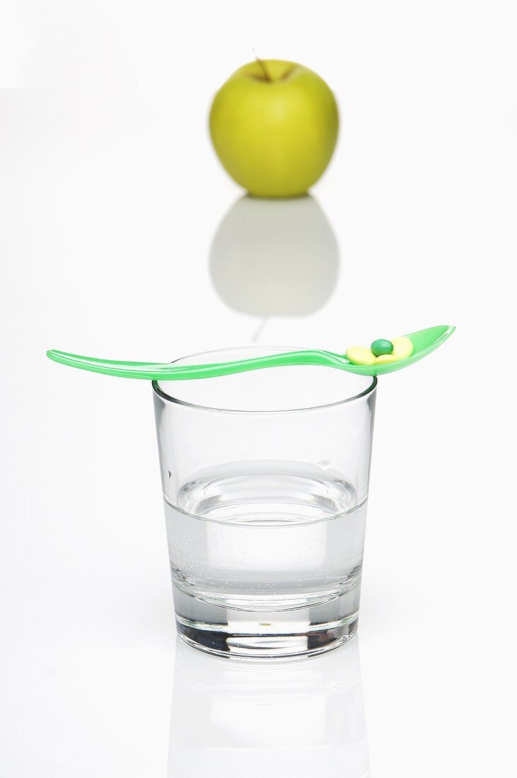 Löffel mit Vitamintabletten auf Wasserglas vor grünem Apfel