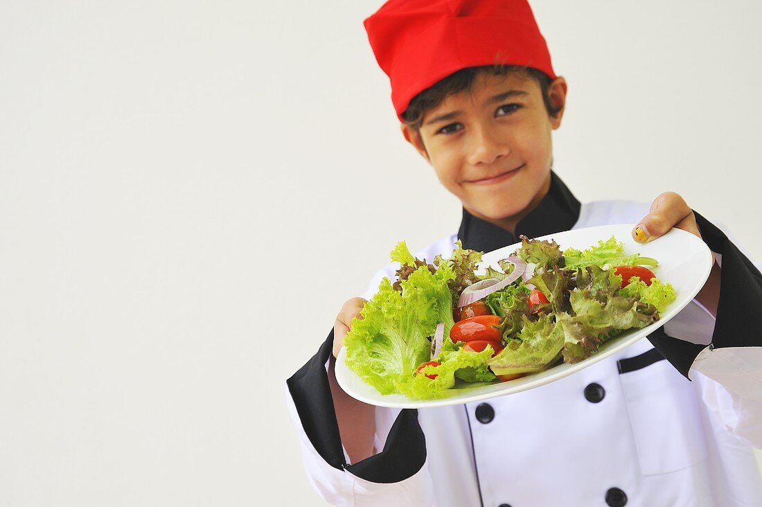 A boy dressed as a chef holding a plate of salad