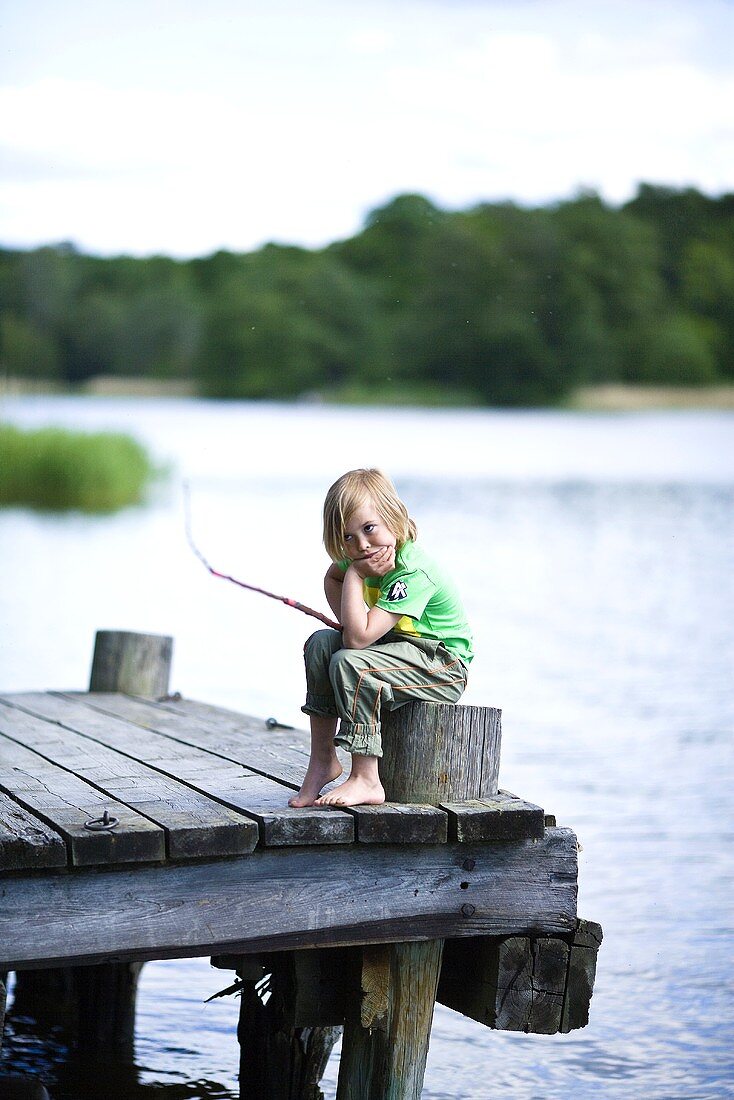 A bored little boy sitting with a fishing rod on a jetty