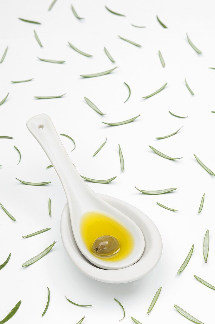 A spoon of olive oil and an olive and scattered rosemary leaves