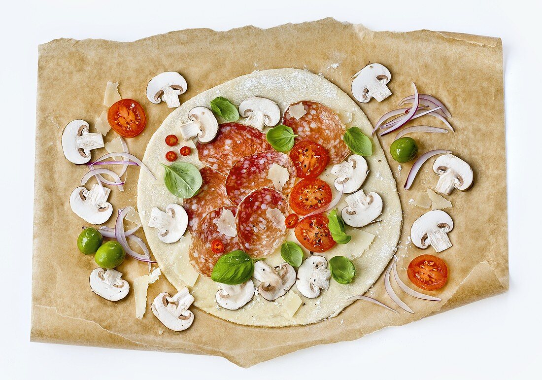 Ungebackene Pizza mit Salami und Champignons auf Backpapier