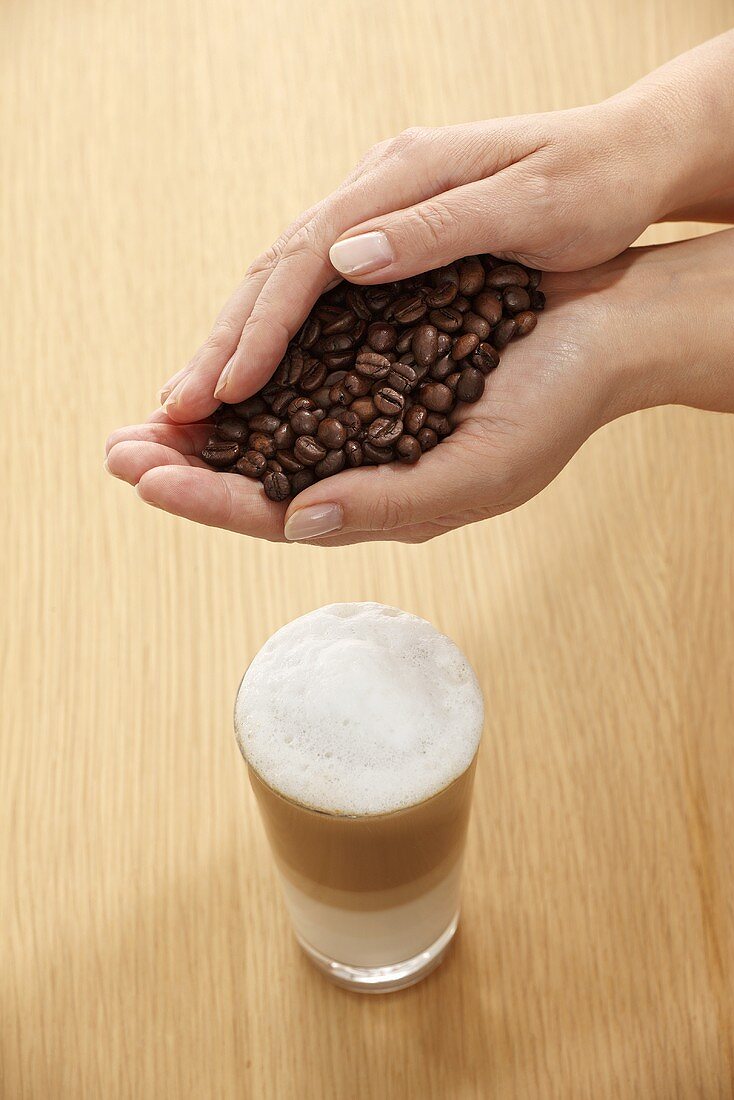 Hände halten Kaffeebohnen über einem Glas Latte Macchiato