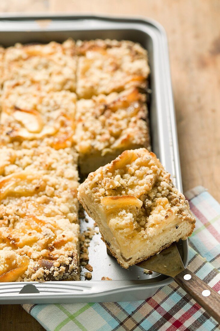 Apple crumble cake in baking tin with piece on server