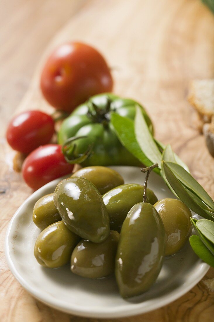 Grüne Oliven auf Teller, dahinter einige Tomaten