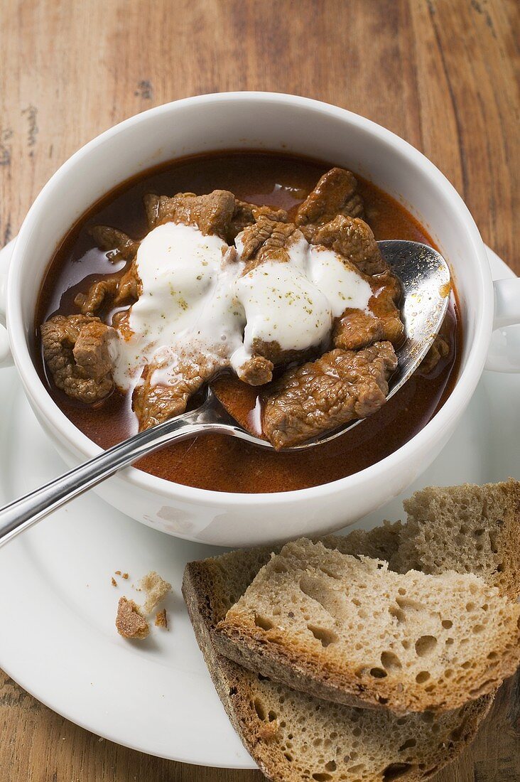 Gulaschsuppe mit saurer Sahne in Suppentasse, Brotscheiben