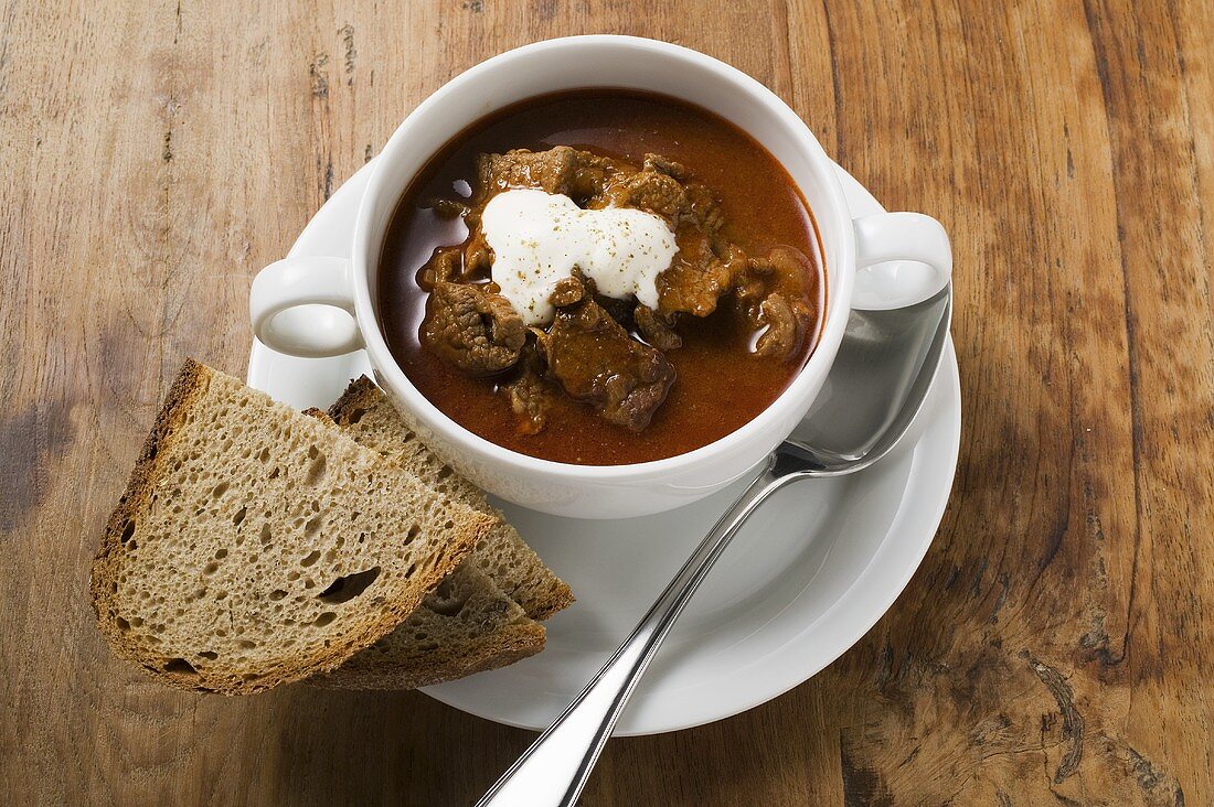 Gulaschsuppe mit saurer Sahne in Suppentasse, Brotscheiben