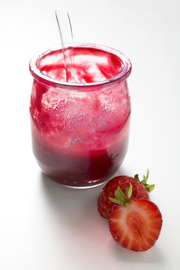 Glas Erdbeermarmelade mit Löffel, daneben frische Erdbeeren