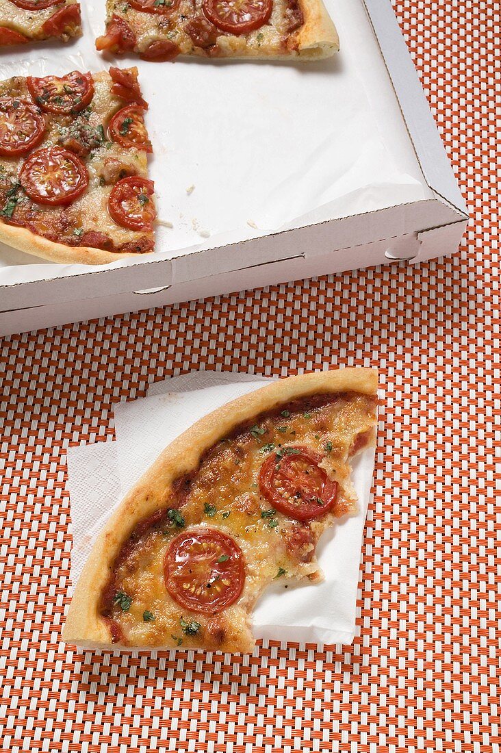 Pizza mit Tomaten im Pizzakarton, daneben angebissenes Stück