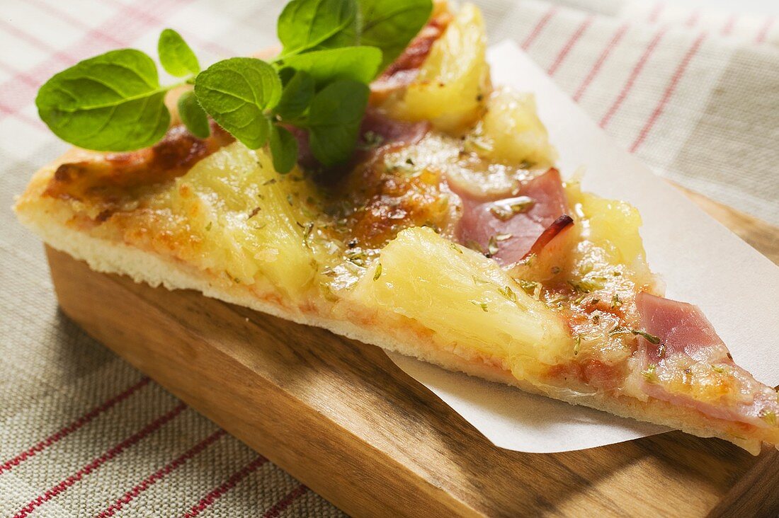 Slice of Hawaiian pizza with fresh oregano on chopping board