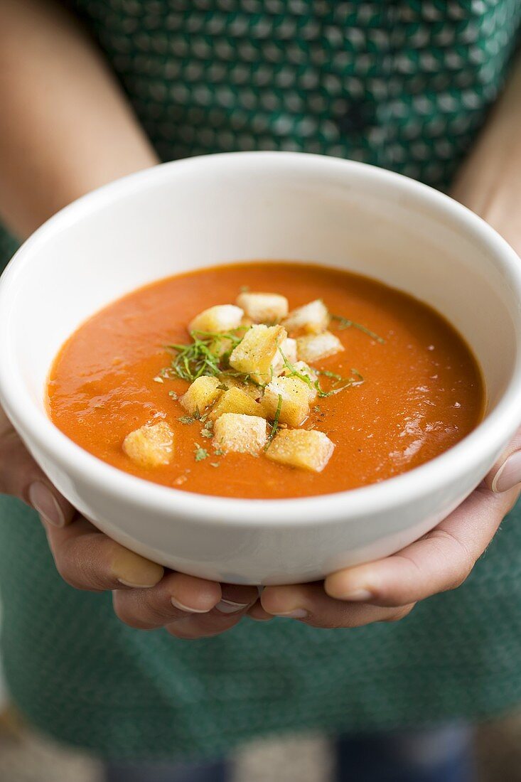 Hands holding bowl of tomato soup with croutons