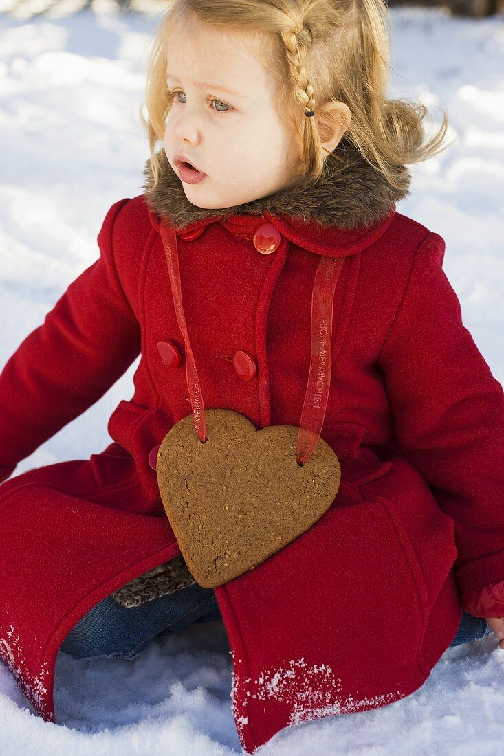 Kleines Mädchen mit Lebkuchenherz sitzt im Schnee